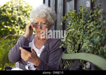 La haute dame assise sur un banc avec ses lunettes de lecture dans son jardin de la lecture de texte message sur son téléphone portable Banque D'Images