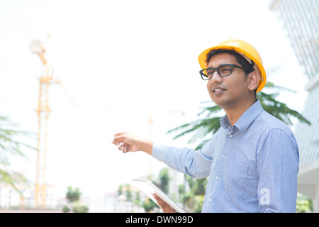 Portrait of a smiling Indian male ingénieur entrepreneur with hard hat pointant vers un site de construction Banque D'Images