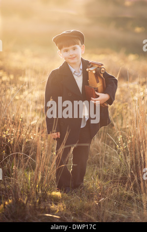 Adorable petit garçon en manteau et cap smiling sur le coucher du soleil sur le terrain, tenant une voiture en bois Banque D'Images