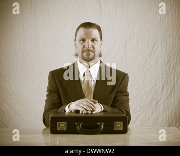 Un 42-year-old woman business man avec une barbiche portant un costume et une cravate est assis à une table de cuisine, les mains croisées sur un attaché-case. Banque D'Images