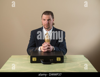 Un 42-year-old woman business man avec une barbiche portant un costume et une cravate est assis à une table de cuisine, les mains croisées sur un attaché-case. Banque D'Images