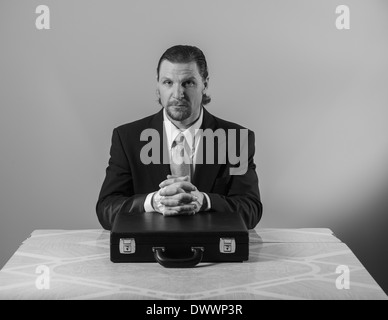 Un 42-year-old woman business man avec une barbiche portant un costume et une cravate est assis à une table de cuisine, les mains croisées sur un attaché-case. Banque D'Images