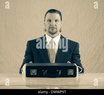 Un 42-year-old woman business man avec une barbiche portant un costume et une cravate est assis à une table de cuisine, les mains croisées sur un attaché-case. Banque D'Images