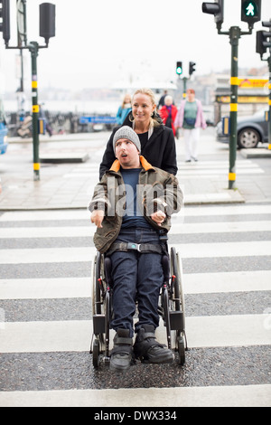 Homme handicapé gardien poussant sur la route en traversant en fauteuil roulant Banque D'Images