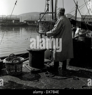 Photo historique Années 1950, d'un pêcheur de mer ou trawlerman en salopette loading paniers de poissons sur le côté du port ou port. Banque D'Images