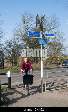 Cycliste femme plus âgée passe un signe sur la National Cycle Route 65 entre York et Beningbrough England UK Mars 2014 Banque D'Images