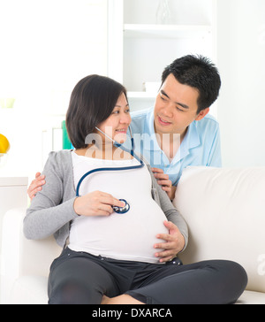 Chinois asiatique pregnant couple vie photo avec salon et canapé à l'écoute de fond ventre stethoscope Banque D'Images