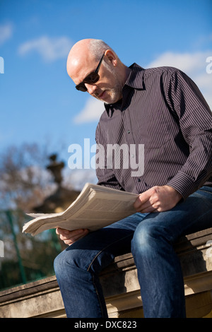 Middle-aged homme chauve assis au soleil en lisant un journal sur un mur de pierre dans un environnement urbain avec sa veste plié à ses côtés Banque D'Images