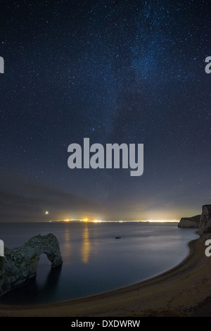 Les stars et Milky Way plus de Durdle Door et la Côte Jurassique, avec les lumières de Weymouth et Portland, Dorset, Angleterre au-delà Banque D'Images