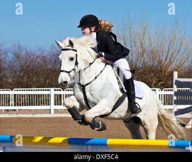 Jeune fille sautant un Cheval poney blanc sur un saut à un événement de saut d'équitation Banque D'Images