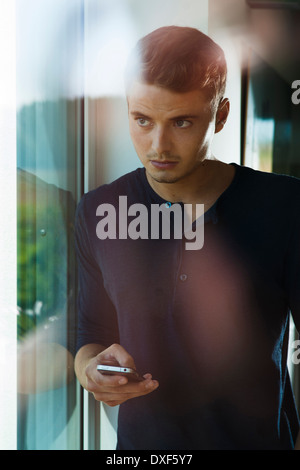 Jeune homme avec Cell Phone looking out Window, Mannehim, Baden-Wurttemberg, Allemagne Banque D'Images