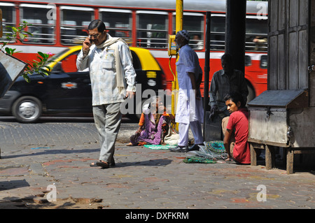 A man talking on a mobile phone passe devant une vieille femme vendant ses marchandises sur un coin de rue à Mumbai Banque D'Images