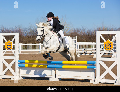 Jeune fille sautant un Cheval poney blanc sur un saut à un événement de saut Banque D'Images