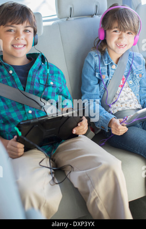 Portrait of happy brother and sister with headphones using digital tablets in back seat of car Banque D'Images