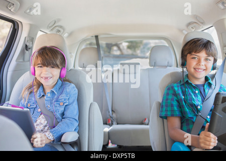 Portrait of happy brother and sister with headphones using digital tablets in back seat of car Banque D'Images