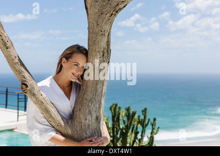 Smiling woman in bathrobe hugging tree in front of ocean Banque D'Images