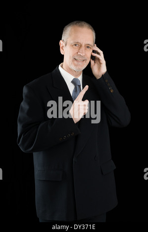 Un homme d'agréable portant un costume noir en souriant tout en parlant au téléphone mobile, sur fond noir Banque D'Images