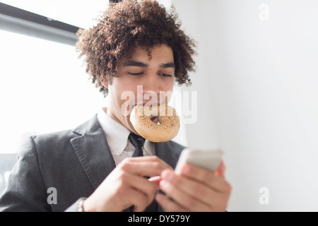 Jeune homme à l'aide de téléphone cellulaire avec bagel en bouche Banque D'Images