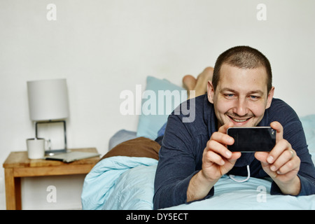 Mid adult man lying on bed taking self portrait on cellphone Banque D'Images