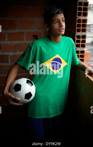 Teen brésilienne graves portant des T-shirt drapeau brésilien est holding football ballon de soccer en regardant par la fenêtre de la favela Banque D'Images