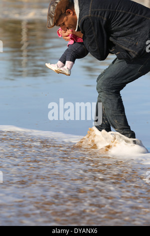 Plus coying Père fille enfant sur une plage de sable humide à Essaouira, Maroc, de ne pas voir une vague venant jusqu'à ce tout dernier min Banque D'Images