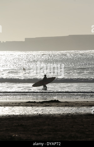 Surfer carrying sa planche de surf le long de la le long du clapotis des vagues d'une plage à Essaouira, Maroc, une fin de soirée Banque D'Images