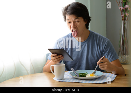 Asian man holding un ipad et qui sort sa langue, d'être stupide, communiquant par ipad/tablette plus prendre le petit déjeuner. Banque D'Images