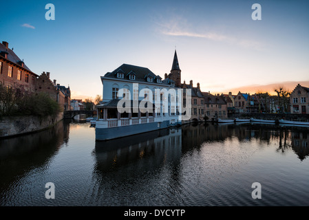 Canaux à Bruges en Belgique | BRUGES, Belgique — Le crépuscule descend sur un canal dans le centre historique de Bruges, où un pavillon de restaurant flottant et des maisons en briques centenaires bordent l'eau près de la Reie. La voie navigable transportait autrefois du trafic marchand qui reliait Bruges à la mer du Nord ; aujourd'hui, ces mêmes canaux attirent des visites en bateau à travers un paysage urbain resté largement inchangé depuis l'ère commerciale médiévale. Banque D'Images