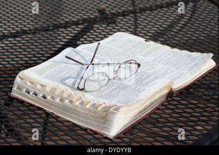 Un gros plan d'une paire de lunettes de lecture reposant sur une portée, Bible ouverte sur une table de patio en fer forgé