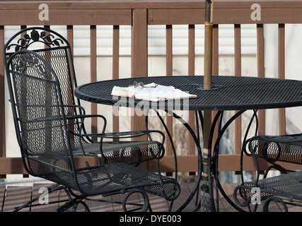 Une paire de lunettes de lecture se trouve au sommet d'une Bible ouverte portant sur une table en fer forgé sur une terrasse en bois avec chaises assorties. Banque D'Images