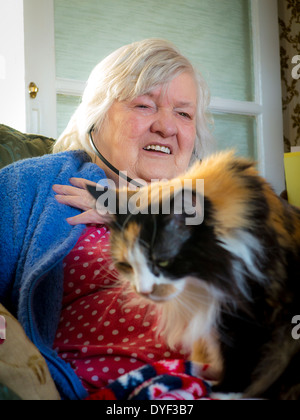 Ancien femme assise dans sa chaise avec son chat sur ses genoux et audio téléphones mobiles dans ses oreilles Banque D'Images