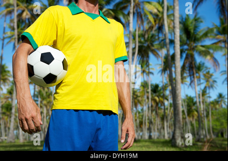 Joueur de football brésilien au Brésil en permanent uniforme couleurs bosquet de palmiers tropicaux holding soccer ball Banque D'Images