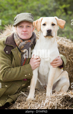 Une femme portant des vêtements de tir assis sur des bottes de paille à l'extérieur avec un chien labrador retriever jaune Banque D'Images