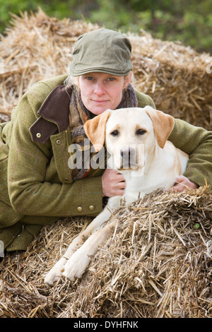 Une femme portant des vêtements de tir assis sur des bottes de paille à l'extérieur avec un chien labrador retriever jaune Banque D'Images