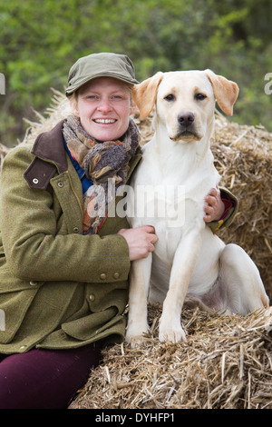 Une femme portant des vêtements de tir assis sur des bottes de paille à l'extérieur avec un chien labrador retriever jaune Banque D'Images