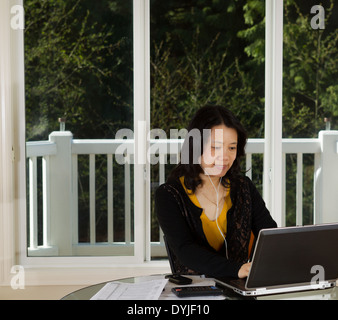 Photo de femme mature à écouter de la musique tout en travaillant à la maison avec un ordinateur portable, téléphone cellulaire, calculatrice et documents sur le dessus du tableau Banque D'Images