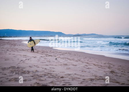 Un surfeur solitaire revient de les vagues sur une plage de surf au crépuscule à Wollongong, Australie. Banque D'Images