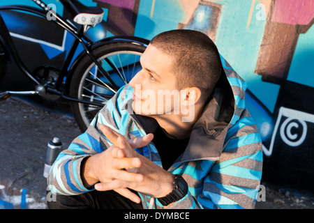 L'artiste graffiti jeune homme assis en face de pièce finie sur mur avec city cruiser bike leaning à côté de lui. La culture urbaine. Banque D'Images