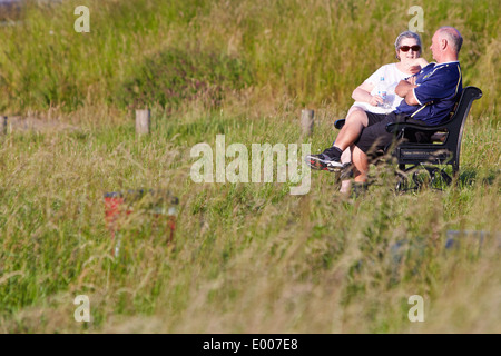 Couple de personnes âgées s'asseoir sur un banc au soleil entouré d'herbe dans Brill village, Buckinghamshire Banque D'Images