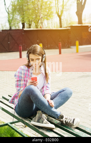 Cute happy young woman in park d'écouter de la musique sur téléphone portable assis sur un banc. Banque D'Images