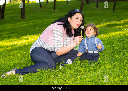 Mère et bébé garçon assis dans l'herbe et en soufflant de l'pissenlit fleur Banque D'Images