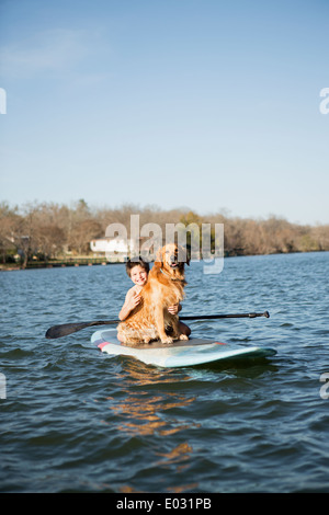 Un enfant et un chien retriever assis sur un paddleboard sur l'eau. Banque D'Images