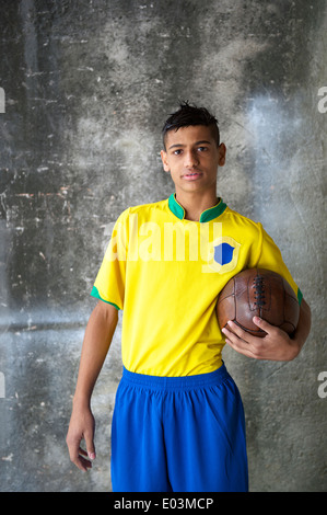 Smiling young Brazilian soccer player en équipe du Brésil de football couleurs holding vintage contre mur béton favela Banque D'Images