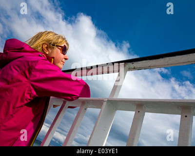 Femme blonde à lunettes et manteau pourpre surplombant la mer depuis le pont d'un navire Banque D'Images