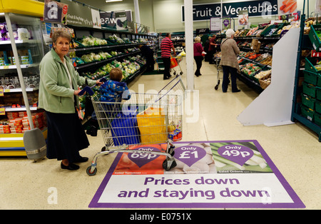 Une vieille dame shopping dans supermarché Tesco, poussant un chariot sur un plancher panneau annonçant des prix bas Banque D'Images