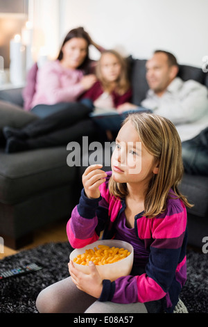 Jeune fille ayant des en-cas tout en regardant la télévision sur le plancher avec la famille en arrière-plan Banque D'Images