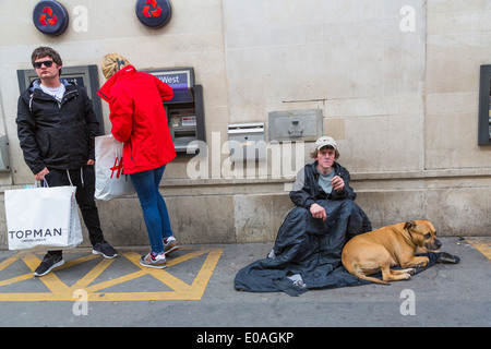 Les jeunes du millénaire retirent de l'argent à un guichet automatique tandis qu'un homme sans domicile et son chien sont assis à proximité en suppliant pour de l'argent, Londres Angleterre Royaume-Uni Banque D'Images