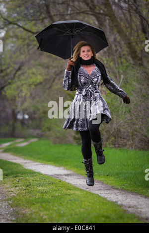 Femme fatale blonde en sautant et smiling holding un parapluie noir sur un chemin de terre Banque D'Images