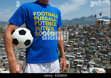 Homme debout dans brésilien de football international t-shirt holding soccer ball en face de taudis favela de Rio de Janeiro d'arrière-plan Banque D'Images