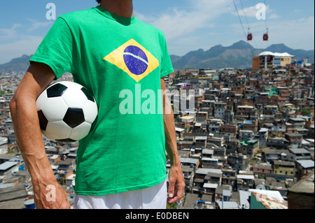 Joueur de football brésilien au Brésil permanent flag t-shirt holding soccer ball en face de taudis favela de Rio de Janeiro d'arrière-plan Banque D'Images